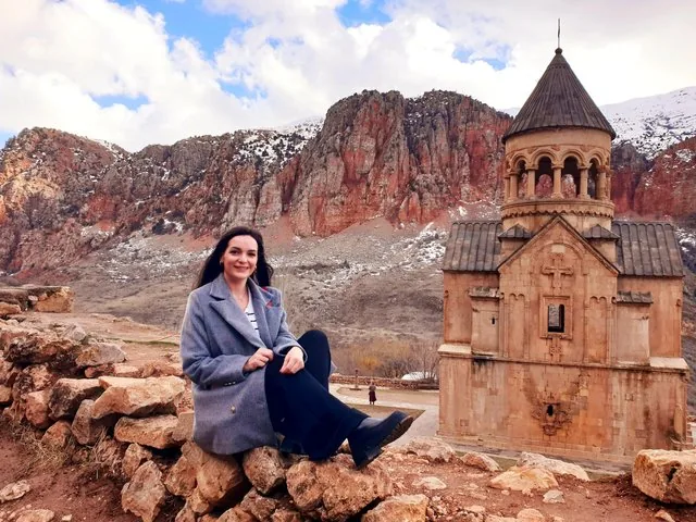 Woman sitting on stone wall with Noravank Monastery and snowy cliffs behind